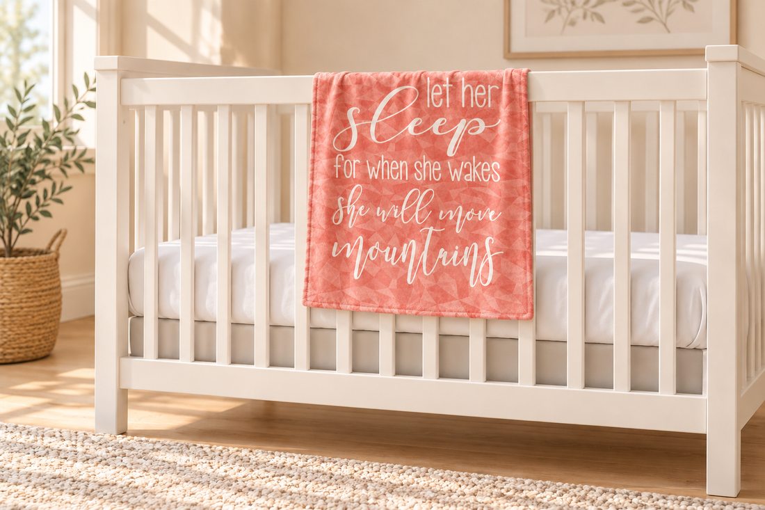 Small pink-and-peach Let Her Sleep baby blanket displayed over the outside rail of an empty white crib.